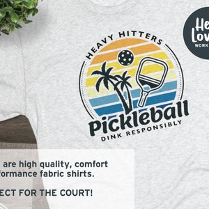 Może przedstawiać: Szary T-shirt z kolorową grafiką przedstawiającą paletkę do pickleballa i zach&oacute;d słońca. Na grafice widnieje tekst "Heavy Hitters" i "Pickleball Dink Responsibly". Tekst "Hello Lovely Workshop" znajduje się w k&oacute;łku w prawym g&oacute;rnym rogu koszulki.