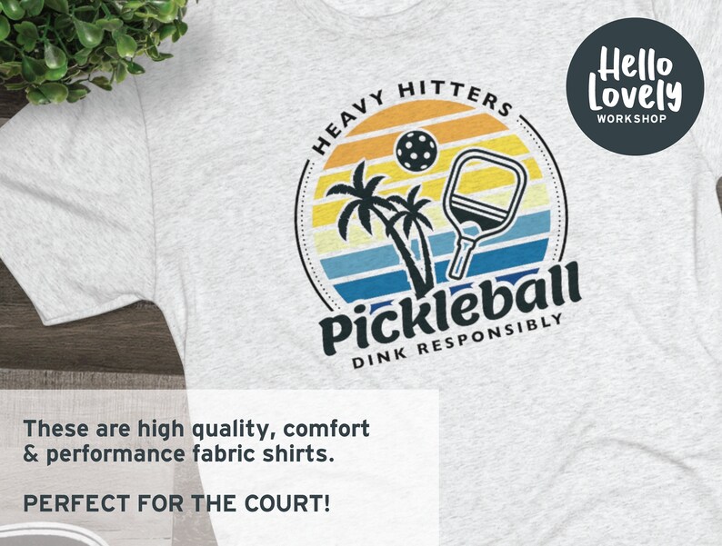 Może przedstawiać: Szary T-shirt z kolorową grafiką przedstawiającą paletkę do pickleballa i zach&oacute;d słońca. Na grafice widnieje tekst "Heavy Hitters" i "Pickleball Dink Responsibly". Tekst "Hello Lovely Workshop" znajduje się w k&oacute;łku w prawym g&oacute;rnym rogu koszulki.