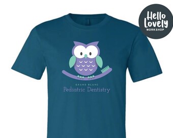 May include: Teal blue t-shirt featuring a purple owl design with mint green accents perched on a toothbrush. The shirt includes the text "Grand Blanc Pediatric Dentistry" in a light teal color. A black circle with white text that says "Hello Lovely Workshop" is in the upper right corner.
