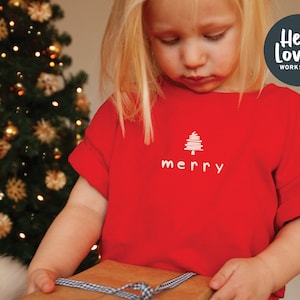 Può includere: Un bambino indossa una maglietta rossa con un albero di Natale ricamato in bianco e la parola "allegro" su di essa. Il bambino tiene una scatola regalo marrone con un nastro a quadri blu e bianco.