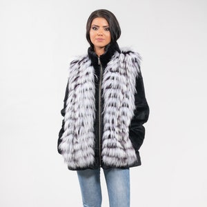 May include: A black fur vest with a white and black striped pattern. The vest is worn over a black shirt and blue jeans.