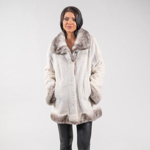 以下が含まれることがあります： 茶色のファーの襟とカフスが付いた白いフェイクファーコート。コートは膝丈で、ボタン留めです。モデルは黒いレギンスと黒いブーツを履いています。