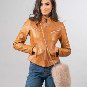 May include: A tan leather jacket with a stand-up collar and zip pockets. The jacket is worn with blue jeans and a light pink fur crossbody bag with a gold chain strap. The model is standing against a grey background.
