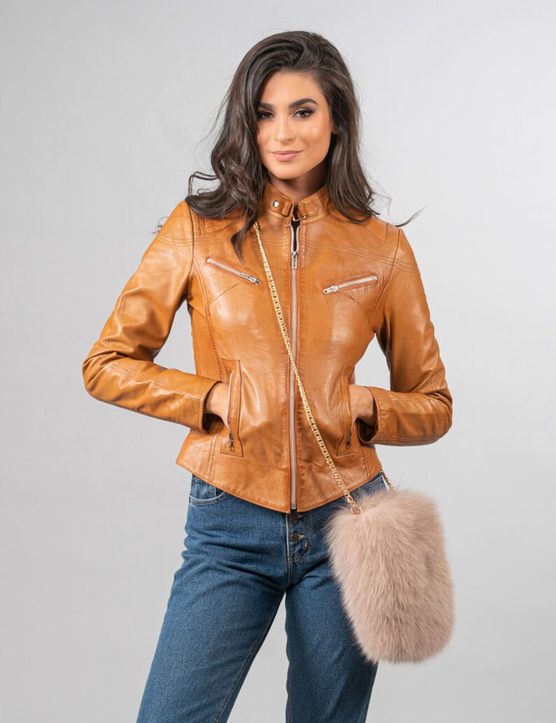 May include: A tan leather jacket with a stand-up collar and zip pockets. The jacket is worn with blue jeans and a light pink fur crossbody bag with a gold chain strap. The model is standing against a grey background.