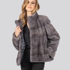 Pode incluir: Um casaco de pele cinzento com gola alta, pain&eacute;is horizontais e mangas compridas. O casaco &eacute; usado sobre uma camisa preta de bot&otilde;es e cal&ccedil;as pretas. O forro interior do casaco &eacute; vis&iacute;vel, exibindo um tecido cinzento escuro com padr&atilde;o.