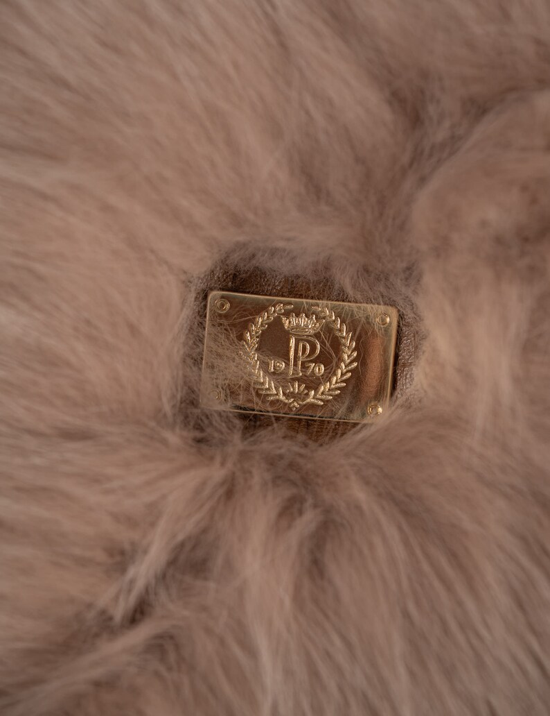 May include: Close-up of a light brown, fluffy material with a rectangular, gold-coloured metal plaque. The plaque features a crown, the letter "P", and the numbers "1970" within a laurel wreath design.