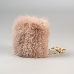 May include: A blush-coloured, rectangular, fluffy handbag with a gold chain strap. The bag is made of soft, textured fur and has a simple, elegant design. The chain is thin and delicate.
