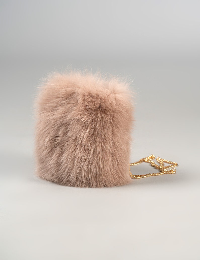 May include: A blush-coloured, rectangular, fluffy handbag with a gold chain strap. The bag is made of soft, textured fur and has a simple, elegant design. The chain is thin and delicate.