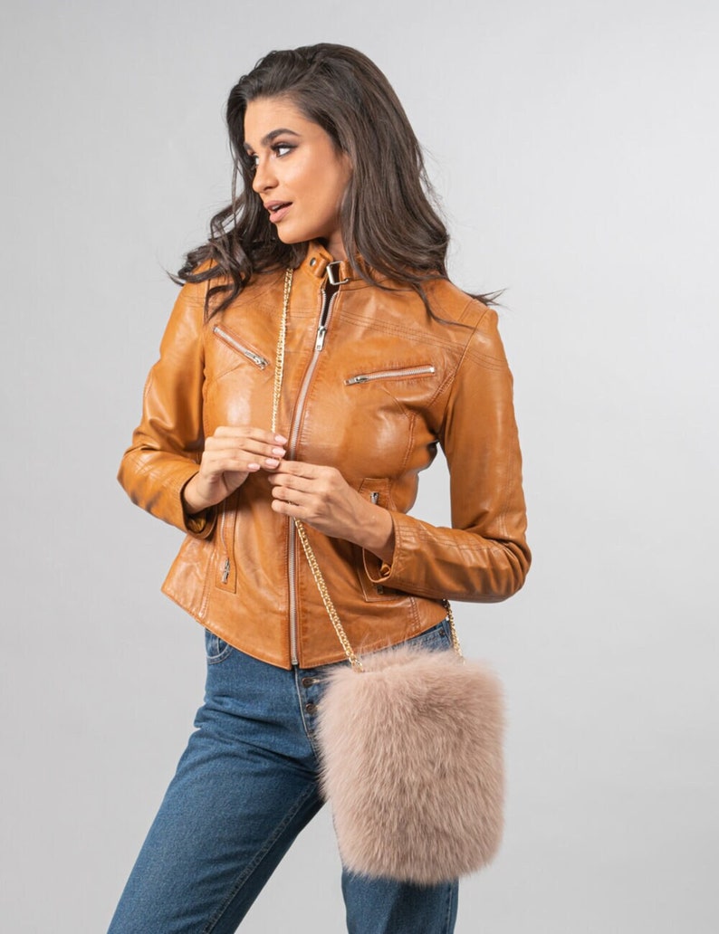 May include: A tan leather jacket with a zip-up front and a gold chain strap purse with a fluffy, light pink exterior. The model is wearing blue jeans.