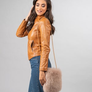 May include: A tan leather jacket with a gold chain strap purse. The purse is made of fluffy, light brown fur. The model is wearing blue jeans.