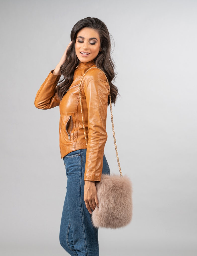 May include: A tan leather jacket with a gold chain strap purse. The purse is made of fluffy, light brown fur. The model is wearing blue jeans.