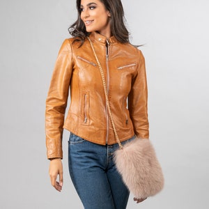 May include: A tan leather jacket with a zip-up front and multiple pockets. The jacket is worn with blue jeans and a light pink fur purse with a gold chain strap. The outfit is set against a neutral background.