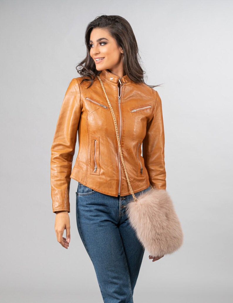 May include: A tan leather jacket with a zip-up front and multiple pockets. The jacket is worn with blue jeans and a light pink fur purse with a gold chain strap. The outfit is set against a neutral background.