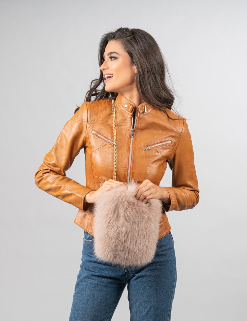 May include: A tan leather jacket with a zip-up front and a small, light pink fluffy purse with a gold chain strap. The jacket has a classic biker style with a stand-up collar and zippered pockets.