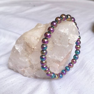 May include: A beaded bracelet with iridescent, multicolored beads. The beads have a metallic sheen, reflecting shades of purple, green, and gold. The bracelet is resting on a white, textured surface, showcasing its vibrant colors and design.