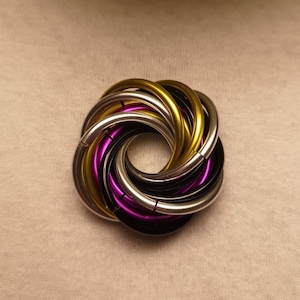 Op de afbeelding: Een veelkleurige metalen ring sculptuur met in elkaar grijpende lussen. De sculptuur heeft ringen in goud, zilver, zwart en paars. Het ontwerp is cirkelvormig, met de ringen die een bloemachtig patroon vormen. De achtergrond is een neutrale beige stof.