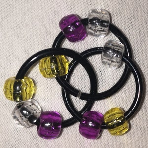 Op de afbeelding: Een zwarte metalen ring met zes gekleurde kralen. De kralen zijn geel, paars en zilver met een glinsterende afwerking.