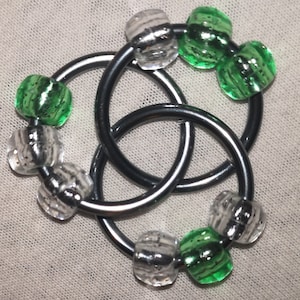 May include: A silver-coloured metal ring knot design with clear and green glass beads. The beads are round and textured. The knot is a three-part design, with each part intertwined. The background is a light beige fabric.