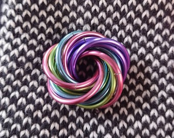 May include: A multi-colored, circular metal object with a rainbow gradient of colors, including pink, purple, green, and blue. The object is set against a black and white knit fabric background.
