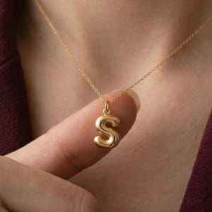 May include: A gold-colored necklace with a pendant shaped like the letter "S". The pendant is a rounded, bubble-style design. The necklace is displayed against a burgundy background.
