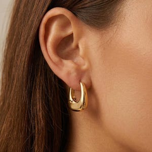 May include: A close-up of a gold-coloured, chunky hoop earring. The earring has a smooth, rounded design and is worn on the ear. The earring is a bold, modern piece of jewellery. The brand name "Foorianco London" is visible in the bottom right corner.