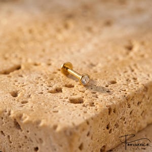 Pode incluir: Uma joia corporal tipo labret em tom dourado com uma gema redonda transparente engastada em bisel na extremidade de uma haste reta com um disco circular plano na parte posterior, sobre uma superfície de pedra bege porosa e texturizada.
