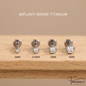Könnte beinhalten: Vier silberfarbene Labret-Stecker aus Implantat-Titan mit klaren, runden Edelsteinen. Die Stecker sind in einer Reihe angeordnet und mit 2mm, 2.5mm, 3mm und 4mm beschriftet, was die Größe der Edelsteinfassung angibt. Text IMPLANT-GRADE TITANIUM.