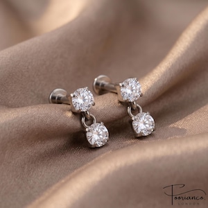 Pode incluir: Um par de brincos labret em tom prateado com haste de base plana e duas pedras de zircônia cúbica transparentes de corte redondo pendentes em engastes de garras. Cada brinco consiste numa pedra superior ligada a uma segunda por um pequeno anel.