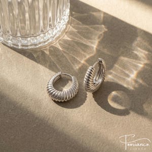 May include: Silver hoop earrings with a ridged design. The earrings are displayed on a neutral surface with shadows and a glass object in the background. The earrings are from the brand Poorianco London.