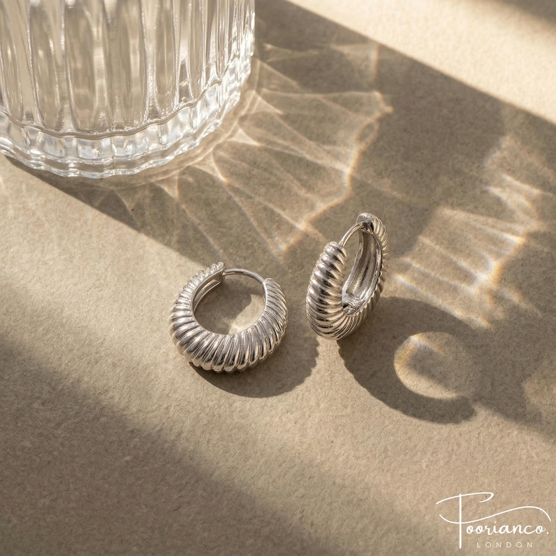 May include: Silver hoop earrings with a ridged design. The earrings are displayed on a neutral surface with shadows and a glass object in the background. The earrings are from the brand Poorianco London.