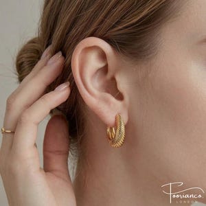May include: A close-up of a gold-coloured hoop earring with a ridged design. The earring is worn on the ear and has a shiny finish. The image shows part of the face and the earring. The earrings are a medium size. The earrings are part of the jewelry category.