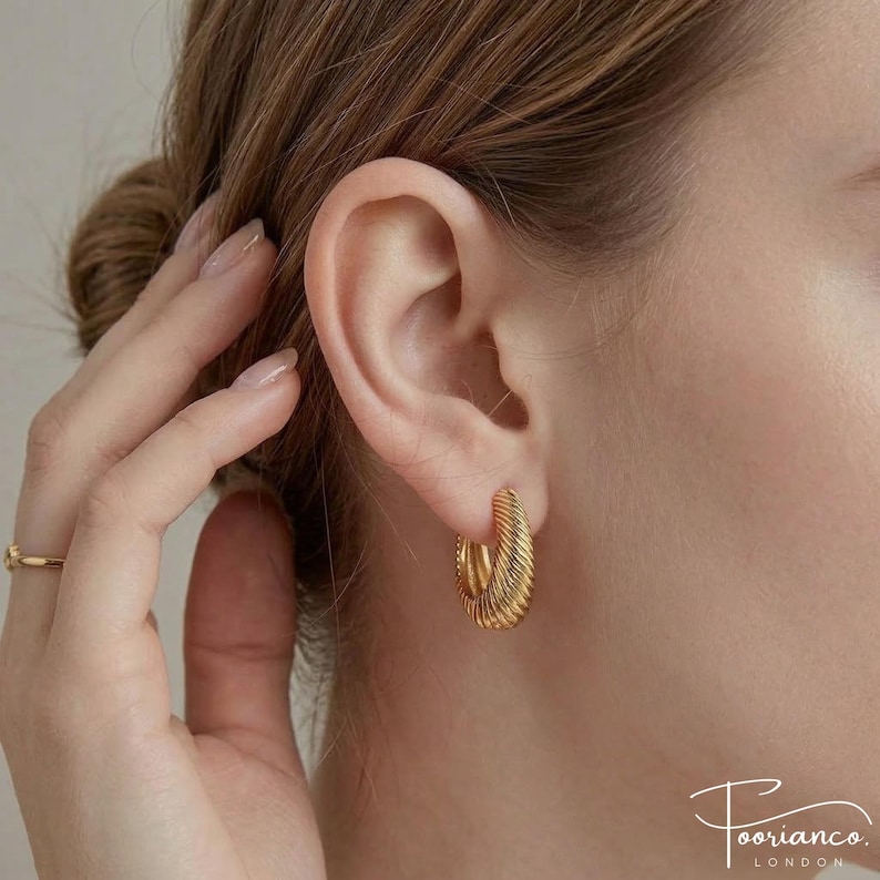 May include: A close-up of a gold-coloured hoop earring with a ridged design. The earring is worn on the ear and has a shiny finish. The image shows part of the face and the earring. The earrings are a medium size. The earrings are part of the jewelry category.