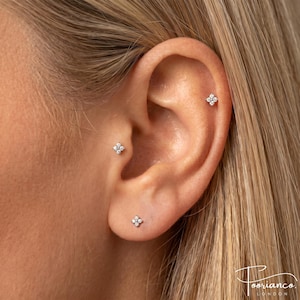 May include: Three silver-toned stud earrings, each featuring a four-petaled flower design set with small clear stones. One stud is placed in the earlobe, one in the tragus, and one in the upper cartilage, all displaying a matching floral motif.