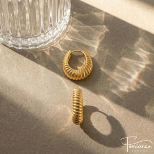 May include: A pair of gold-coloured hoop earrings with a ribbed design. The earrings are displayed on a neutral surface with a glass object in the background. The earrings have a polished finish and a classic, elegant style.