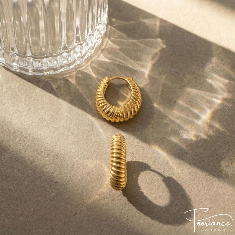 May include: A pair of gold-coloured hoop earrings with a ribbed design. The earrings are displayed on a neutral surface with a glass object in the background. The earrings have a polished finish and a classic, elegant style.
