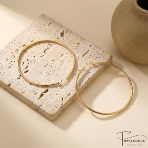 May include: A pair of large, thin, circular gold hoop earrings with a smooth, polished finish. Each earring features a simple hinged post closure mechanism. The earrings are displayed as a matching set, showcasing their uniform round shape.
