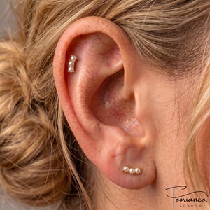 May include: A gold-toned ear piercing jewellery set consisting of two separate pieces, each featuring three small, round white pearls arranged in a linear row. One piece is positioned in the upper cartilage, while the other is in the earlobe.