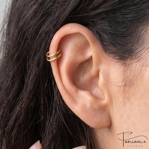 May include: A gold-toned double hoop ear cuff worn on the upper cartilage of the ear. The jewellery features two thin, smooth, circular bands that sit parallel to each other, creating a layered appearance on the outer edge of the ear structure.