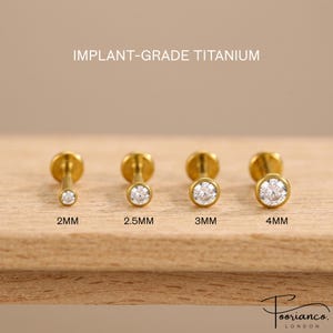 May include: Four gold-tone flat back stud earrings with clear round stones, labelled IMPLANT-GRADE TITANIUM. The studs are arranged by size: 2mm, 2.5mm, 3mm, and 4mm. Each earring features a circular bezel setting holding a single faceted stone.