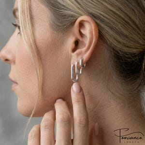 May include: A person's ear adorned with three silver hoop earrings of varying sizes. The earrings have a rectangular design with rounded corners. The person has fair skin and blonde hair. The earrings are the central element of the image.