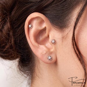 Peut inclure: Trois boucles d'oreilles en forme de fleur de couleur argentée avec une pierre centrale transparente, portées au lobe, au tragus et à l'hélix. Chaque boucle présente un design à cinq pétales avec une finition métallique et une pierre centrale.