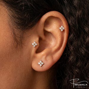 Könnte beinhalten: Drei silberfarbene Ohrstecker, jeder mit einem vierblättrigen Blumendesign aus kleinen, klaren, rund geschliffenen Steinen. Die Ohrstecker sind am Ohrläppchen, am Tragus und am oberen Knorpel platziert und bilden ein abgestimmtes Set.