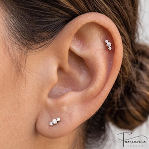 Può includere: Un set di gioielli per piercing all'orecchio color argento con due orecchini a lobo abbinati, ciascuno composto da tre piccole pietre trasparenti a taglio rotondo disposte in linea. Un orecchino è sul lobo e l'altro sulla cartilagine.