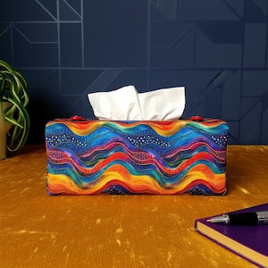 May include: A rectangular fabric tissue box cover featuring a vibrant, multi-coloured wave pattern in shades of blue, purple, orange, and yellow, accented with small white dots. The cover has two small red buttons on the top edge and holds white tissues.