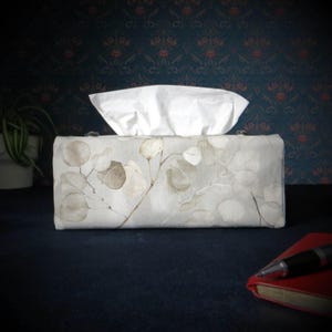 May include: A white tissue box cover with a floral design featuring light brown leaves and branches on a light grey background.