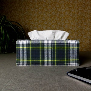 May include: A tissue box cover with a plaid pattern in shades of green, blue, white, and yellow. The tissue box is sitting on a grey surface.