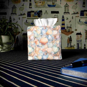 May include: A square tissue box cover decorated with a seashell design in blue, beige, and orange. White tissues protrude from the top. It rests on a striped surface, with coastal-themed wallpaper in the background.