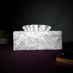 May include: A white tissue box cover with a floral design featuring grey roses. The cover is made of fabric and has a rectangular shape.