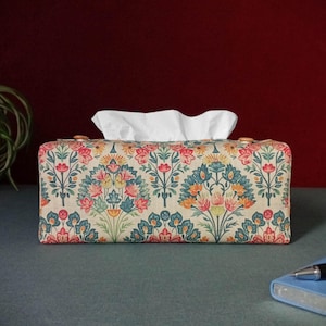 May include: A rectangular tissue box cover with a floral design in red, blue, yellow, and green. The fabric cover has a beige background. White tissues are emerging from the top.