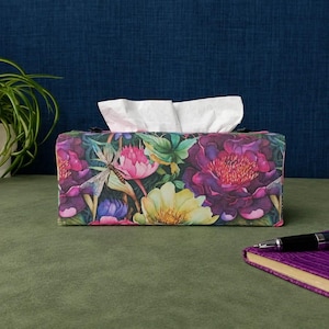May include: A rectangular tissue box cover featuring a vibrant floral pattern with large purple, pink, and yellow flowers, green leaves, and a dragonfly. White tissues protrude from the top opening of the box, which sits on a flat surface.